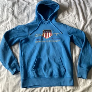 Blå hoodie från GANT - Säljer en snygg blå hoodie från GANT med broderad logga och text 'American Sportswear' på framsidan. Tröjan har en klassisk känguruficka och justerbar huva med dragsko. Perfekt för en avslappnad stil!