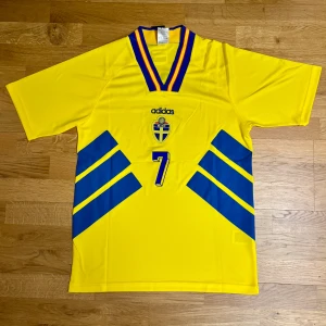 Sverige 1994 fotbollströja Larsson - Säljer en gul fotbollströja från Adidas med blå detaljer och nummer 7 på framsidan. Tröjan har korta ärmar och ett märke från Svenska Fotbollförbundet. Perfekt för fotbollsfans!