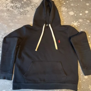 Ralph lauren hoodie  - Säljer en stilren svart hoodie från Ralph Lauren med en liten röd logga på bröstet. Den har en klassisk känguruficka och vita snören i huvan. Perfekt för en avslappnad look.