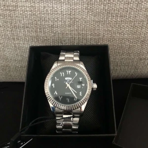 Silverfärgad klocka med svart urtavla - Elegant silverfärgad klocka med svart urtavla och arabiska siffror. Klockan har en kvartsrörelse och en robust metallarmband. Perfekt för den som söker en stilren accessoar.