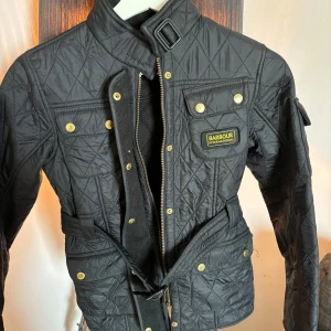 Barbour jacka  - Snygg svart quiltad jacka från Barbour med guldknappar och dragkedja. Jackan har en hög krage med spänne och flera praktiska fickor. Perfekt för en stilren look.