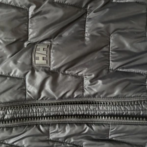 Svart quiltad jacka från Helly Hansen - Snygg svart quiltad jacka från Helly Hansen med dragkedja framtill. Perfekt för kyligare dagar med sin stilrena design och bekväma passform. Jackan har långa ärmar och en diskret logga på bröstet.