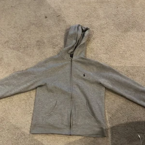 Grå hoodie från Polo Ralph Lauren - Säljer en stilren grå hoodie från Polo Ralph Lauren. Den har en dragkedja framtill och en klassisk huva. Perfekt för en avslappnad look. 🐎