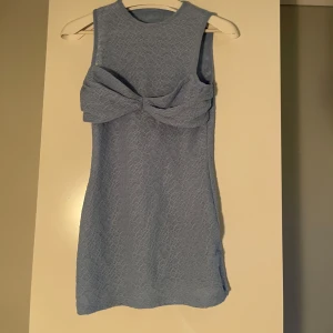 Ljusblå Klänning  - Snygg ljusblå klänning med en stor rosett framtill. Klänningen är ärmlös och har en kort längd, perfekt för varma dagar. Materialet har en subtil textur som ger en extra touch.