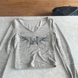 Grå croppad långärmad tröja - Grå/beige långärmad croppad tröja med v-ringning och tryck av döskalle och svarta detaljer framtill. Mjuk och tunn, perfekt för lager-på-lager. Snygg till både jeans och kjol för en edgy vibe.