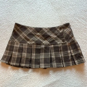 Pleated minikjol - Supersöt brun och beige rutig minikjol med plisserad nederdel och tre knappar framtill. Kjolen har inbyggda shorts under och dragkedja i sidan för enkel påklädning. Perfekt för en trendig och preppy look! 