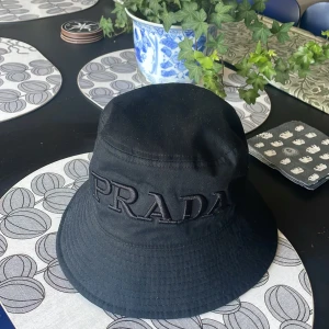 Svart bucket hat från Prada - Snygg svart bucket hat från Prada med broderad logga framtill. Klassisk form med bred brätte och diskret ton-i-ton brodyr. Tillverkad i bomull, perfekt för att lyfta din streetwear-look.
