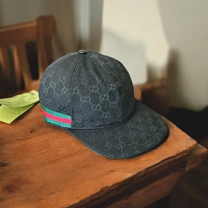 Gucci svart keps med GG-mönster - Svart keps från Gucci med klassiskt GG-monogram över hela ytan och grönt/rött band på sidan. Justerbar rem baktill i skinn och böjd skärm. Snygg och stilren accessoar som lyfter din outfit.