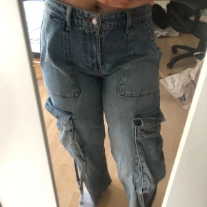 Blå cargojeans med vida ben från Zara - Säljer ett par blå cargopants i jeansmaterial med bred passform och stora fickor på sidorna. Byxorna har hög midja, råa kanter nertill och dragkedjedetaljer vid bensluten. Perfekta för en avslappnad och trendig look.