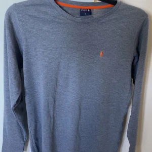 Grå långärmad tröja Polo Ralph Lauren - Snygg grå långärmad tröja från Polo Ralph Lauren i bomull. Klassisk passform med rund halsringning och orange broderad logga på bröstet. Perfekt basic-plagg med stilren look och diskreta detaljer. PRIS KAN DISKUTERAS VID SNABB AFFÄR!