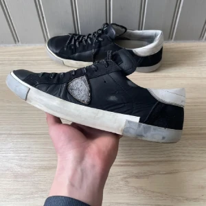 Philippe Model svarta - Svarta sneakers från Philippe Model i storlek 43 med vit häl och grå detalj på sidan. Skorna har snörning med logotyp på skosnörena, platt sula och rund tå. Tillverkade i skinn med detaljer i mocka och syntet. Snygg kontrast mellan svart och vitt, perfekt för en trendig streetstil.