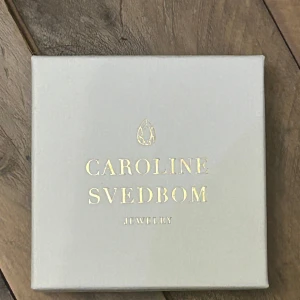 Guldarmband Caroline Svedbom med kristaller - Snyggt armband från Caroline Svedbom i guldtonad metall med tvinnad design. Armbandet har två stora, droppformade kristaller i persikofärg som fångar ljuset superfint. Perfekt statement-piece för dig som gillar att sticka ut.