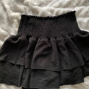 Svart volangkjol från Skue - Supersöt svart kortkjol från Skue med bred smock i midjan och flera lager volanger.