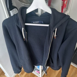 Mörk blå hoodie med dragkedja från Morris - Svart hoodie från Morris i storlek S med vit snörning i huvan och dragkedja framtill. Klassisk modell med fickor och liten broderad logga på bröstet. Perfekt för chill dagar och enkel att matcha med jeans eller joggers.