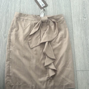 Beige kjol med volang från Vila - Supersnygg beige kortkjol från Vila med volangdetalj framtill och knyt i midjan. Kjolen har en stilren look och stängs med dragkedja bak. Perfekt för dig som gillar en feminin och chic stil.