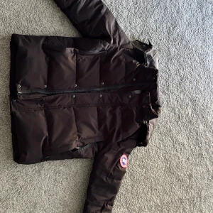Svart dunjacka från Canada Goose - Säljer en svart dunjacka från Canada Goose med klassisk patch på ärmen. Jackan har lång ärm, dragkedja och tryckknappar framtill. Fylld med dun och fjädrar för extra värme, perfekt för kalla dagar. Snygg och tidlös design som funkar till allt. Storlek M PS pälsen fattas men kan användas med stil endå