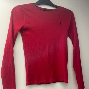 Röd långärmad tröja från Ralph Lauren - Röd långärmad tröja från Ralph Lauren med klassisk rund halsringning och mörkblå broderad logga på bröstet. Tröjan är i mjuk bomull och har en slim passform som sitter snyggt på kroppen. Perfekt för dig som gillar stilrena basics.