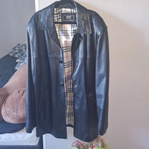 Svart skinnjacka från Kent XL - Stilren svart skinnjacka från Kent i storlek XL. Jackan har klassisk krage, knäppning framtill och två fickor. Insidan är fodrad med rutigt tyg i vitt, blått och rött. Perfekt för dig som gillar tidlös och clean look.