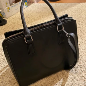 Svart väska chiquelle - Svart väska i skinnimmitation med både handtag och avtagbar axelrem. Väskan har flera fack, dragkedja upptill och en extra ficka med dragkedja på baksidan. Perfekt för laptop och allt du behöver till plugget eller jobbet. Säljer pga att jag inte använder den längre.
