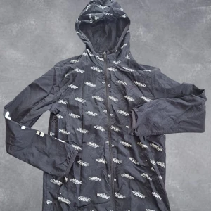 Adidas vindjacka - Svart vindjacka från Adidas med hel dragkedja och huva. Jackan har ett all-over print med vita Adidas-loggor och elastiska muddar vid ärmsluten. Perfekt för dig som gillar sportig streetstyle och vill sticka ut med en snygg loggodesign.