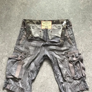 Randiga cargoshorts från Alpha Industries - Snygga cargoshorts från Alpha Industries i grått med tunna svarta ränder. Shortsen har flera fickor, coola detaljer som knappar, bälteshällor och förstärkta sömmar. Perfekta för dig som gillar en streetig och funktionell stil. Materialet känns slitstarkt och är troligtvis bomull.