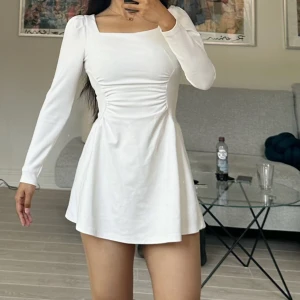 Vit långärmad kortklänning  - Supersöt vit kortklänning från NAKD med långa ärmar och fyrkantig ringning. Klänningen har snygg rynkdetalj i midjan som ger en smickrande siluett. Tillverkad i mjuk bomull, perfekt för vår och sommar. Dragkedja bak för enkel påklädning.