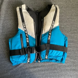 Blå flytväst från Baltic - Flytväst från Baltic i blått och vitt med svarta detaljer. Västen har dragkedja framtill, justerbart midjebälte och svarta remmar med Baltic-logga. Materialet är syntet och designen är sportig och funktionell, perfekt för vattenaktiviteter.