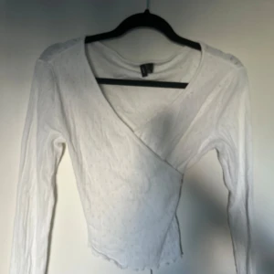 Vit omlottopp med knyt topp - Supersöt vit omlottopp från veromoda med långa ärmar och knyt i midjan. Tunt ribbat material med små hålmönster och vågig kant nedtill. Perfekt för dig som gillar en feminin och trendig stil. Sitter snyggt och är lätt att matcha med jeans eller kjol.