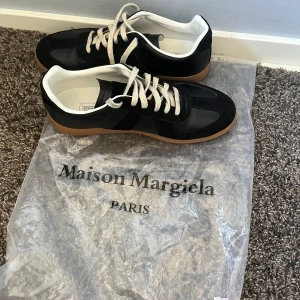 Svarta Maison Margiela sneakers med mockadetaljer - Svarta sneakers från Maison Margiela med snygga detaljer i mocka och skinn, vita snören och vit insida. Skorna har en klassisk gummisula i brunt och en stilren, tidlös design som passar till det mesta. Perfekta för dig som gillar minimalistisk och clean stil.