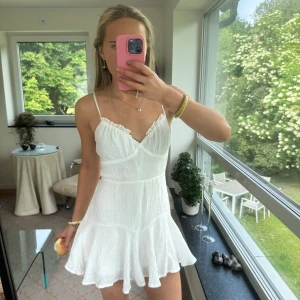 Vit somrig kortklänning med volang - Superfin vit kortklänning med tunna axelband och v-ringning. Klänningen har volangkant nertill och lätt rynk vid bysten för en söt look. Perfekt för varma dagar och har en luftig känsla tack vare det tunna bomullstyget.