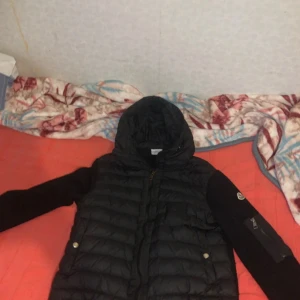 Svart Moncler pufferjacka med huva - Svart pufferjacka från Moncler med stickade ärmar och huva. Jackan har dragkedja framtill, två fickor med tryckknappar och en extra ficka med dragkedja på ärmen. Klassisk Moncler-logga på vänster ärm. Perfekt för kyliga dagar.