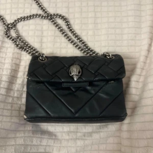 Svart quiltad axelväska från Kurt Geiger - Snygg svart axelväska från Kurt Geiger i quiltat skinn med silverfärgad kedja och ikoniskt örnhuvud på locket. Väskan har magnetlås och stilren, boxig form. Perfekt för dig som gillar edgy detaljer och klassisk design.