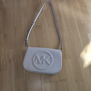 Vit axelväska Michael Kors Brynn - Säljer en stilren axelväska från Michael Kors, modell Brynn. Väskan är i mjukt skinn med en ljurosa ton och har ett stort MK-logo broderat på framsidan. Justerbar axelrem och flera fack inuti för dina essentials. Perfekt för dig som gillar clean och lyxig vibe.