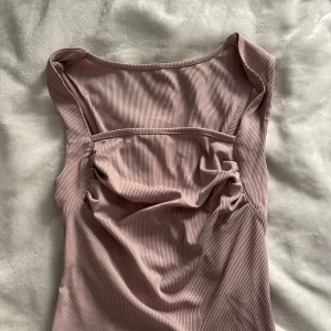 Ribbad ljusrosa linnetopp - Säljer en ribbad ljusrosa linnetopp med breda axelband och snygg rynkdetalj framtill. Toppen är ärmlös och har en mjuk, stretchig känsla som gör den superbekväm. Perfekt att styla med jeans eller kjol för en trendig look.