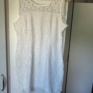 Vit spetsklänning med volangdetalj - Superfin vit kortklänning i spets med blommigt mönster och volangdetaljer vid axlar och nederkant. Ärmlös modell med rund halsringning, perfekt för sommarens alla tillfällen. Söt och stilren look som passar dig som gillar romantiska vibes.