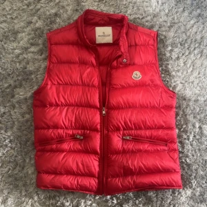 Röd dunväst från Moncler - Riktigt snygg röd dunväst från Moncler med klassisk logga på bröstet. Västen har två dragkedjefickor framtill och quiltad design som ger en puffig look. Perfekt att slänga över en hoodie för extra värme. Stängs med dragkedja hela vägen upp.