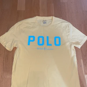 Gul t-shirt från Polo Ralph Lauren - Gul t-shirt från Polo Ralph Lauren med stor blå POLO-logga framtill. Slim fit-modell i mjuk bomull, perfekt för dig som gillar klassisk men ändå sportig stil. Rund halsringning och korta ärmar. Snygg och enkel att matcha med jeans eller shorts.