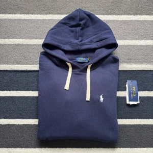 Blå hoodie från Polo Ralph Lauren - Snygg marinblå hoodie från Polo Ralph Lauren med klassisk vit broderad logga på bröstet. Tröjan har huva med vita snören och en stor magficka. Tillverkad i mjuk bomull, perfekt för chill dagar. Långärmad och med ribbade muddar för extra komfort.