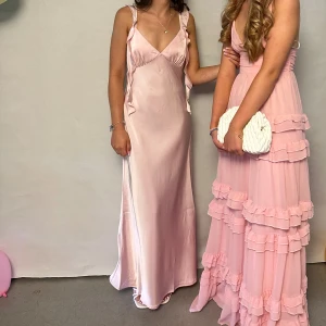 Ljusrosa långklänning i satin - Supersnygg ljusrosa långklänning i satinliknande material med smala axelband och v-ringning. Klänningen har volangdetaljer längs sidorna och en elegant, figurnära passform som faller snyggt längs kroppen. Perfekt för bal eller student! Xs-s