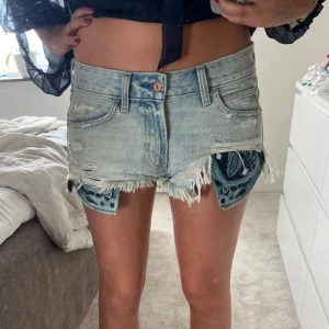Ljusblå slitna jeansshorts med fransar - Säljer ett par ljusblå jeansshorts med råa fransade kanter och slitningar framtill. Shortsen har låg midja, klassiska fickor och coola blå mönstrade detaljer som sticker ut under den slitna kanten. Perfekta för sommaren och en avslappnad streetstil.