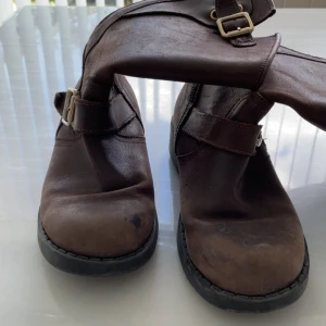 Bruna johnny bulls  - Snygga bruna johnny bulls  boots i skinn med robust känsla. De har dekorativa spännen i mässing och dragkedja baktill för enkel påtagning. Perfekta för dig som gillar en klassisk och tuff stil. Storlek 36 men passar 37