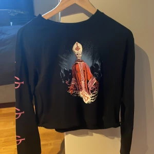 Svart croppad tröja med tryck Ghost - Cool svart croppad tröja från Ghost med stort färgstarkt tryck av Papa Emeritus på bröstet och rosa symboler längs ärmarna. Långärmad modell med ribbade muddar och relaxed fit. Perfekt för dig som gillar bandmerch och vill sticka ut.
