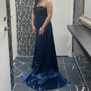 Marinblå axelbandslös balklänning - Säljer en marinblå långklänning med axelbandslös design och glittrig överdel. Klänningen har draperad midja och ett elegant släp. Materialet är glansigt och känns lyxigt, perfekt för bal eller fest. Sitter snyggt och ger en riktigt glam känsla.