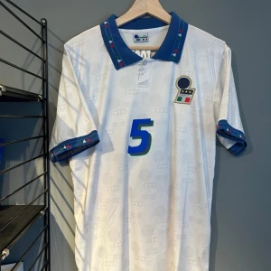 Italien fotbollströja Maldini #5 - Säljer en klassisk vit Italien fotbollströja från Diadora med blå detaljer och nummer 5 på bröstet och ryggen. Kragen och ärmsluten har blått mönster med små färgdetaljer. På ryggen står det 'Maldini'. Perfekt för samlare eller fans!
