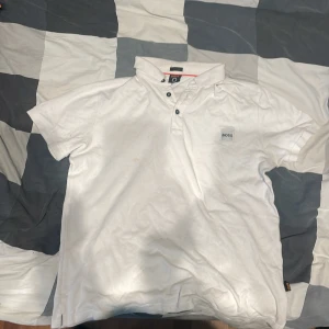 Vit kortärmad t-shirt från BOSS - Vit t-shirt från BOSS med klassisk krage och knappslå framtill. T-shirten har korta ärmar och en liten BOSS-logga på bröstet samt etikett i sidan. Tillverkad i mjuk bomull, perfekt för en clean och stilren look.