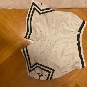 Vita Jordan basketshorts med svarta detaljer - Snygga vita Jordan basketshorts med svarta ränder längs benen och klassisk Jumpman-logga. Shortsen är tillverkade i luftig mesh med snörning i midjan och elastisk linning. Perfekta för sport eller chill, riktigt clean look.