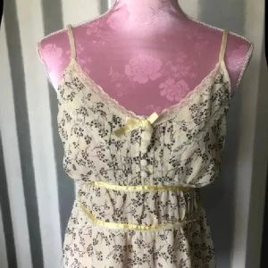 Blommig linne med spetsdetaljer - Supersöt linne i ljusbeige med småblommigt mönster och tunna axelband.linnet har spetskant vid urringningen, dekorativ rosett framtill och gula band i midjan. Perfekt för sommarens varma dagar och har en romantisk vibe.