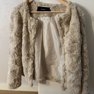 Beige teddyjacka från Vero Moda - Supermjuk och fluffig teddyjacka från Vero Moda i beige färg. Jackan har rund halsringning, långa ärmar och stängs med tryckknappar framtill. Perfekt för dig som vill ha en mysig och trendig look under kyliga dagar.