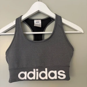 Grå sportig sporttopp från Adidas - Snygg grå linnetopp från Adidas med stort vitt logotryck framtill. Modellen är ärmlös med brottarrygg och tillverkad i ett stretchigt material som passar perfekt för träning. Enkel och stilren design som ger sportig vibe. Använd max 5 gånger 