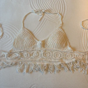 Vit virkad croppad linnetopp - Supersöt vit virkad linnetopp med knyt i nacken och fransar längs nederkanten. Toppen är croppad och har ett boho-inspirerat mönster, perfekt för varma dagar och festivaler.
